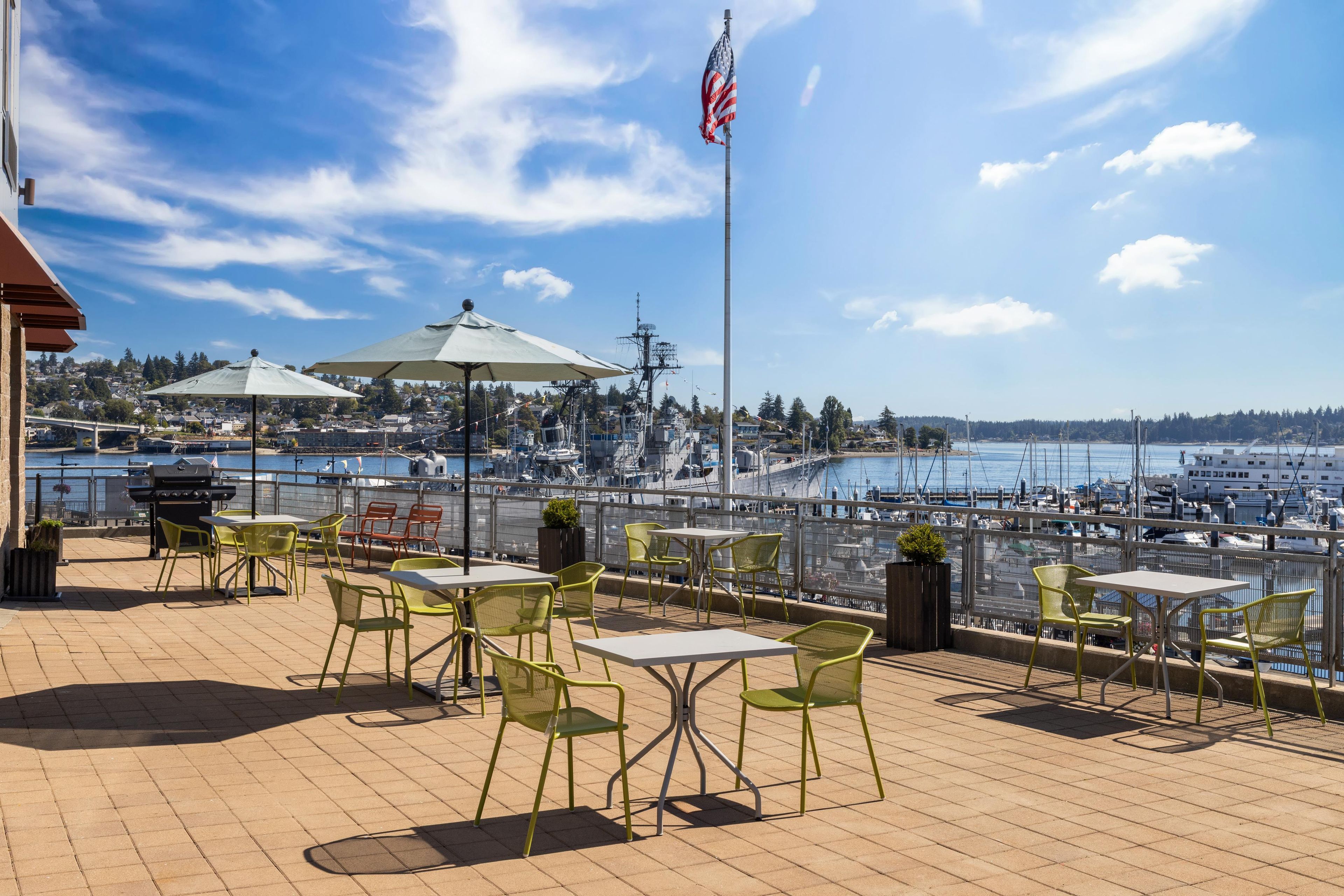 An outdoor terrace with tables and chairs, overlooking a marina with boats and a cityscape in the background.