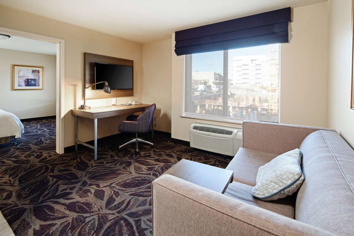A hotel room with a bed, a desk with a chair, a television, a window with a view of buildings, and a sofa with a pillow.