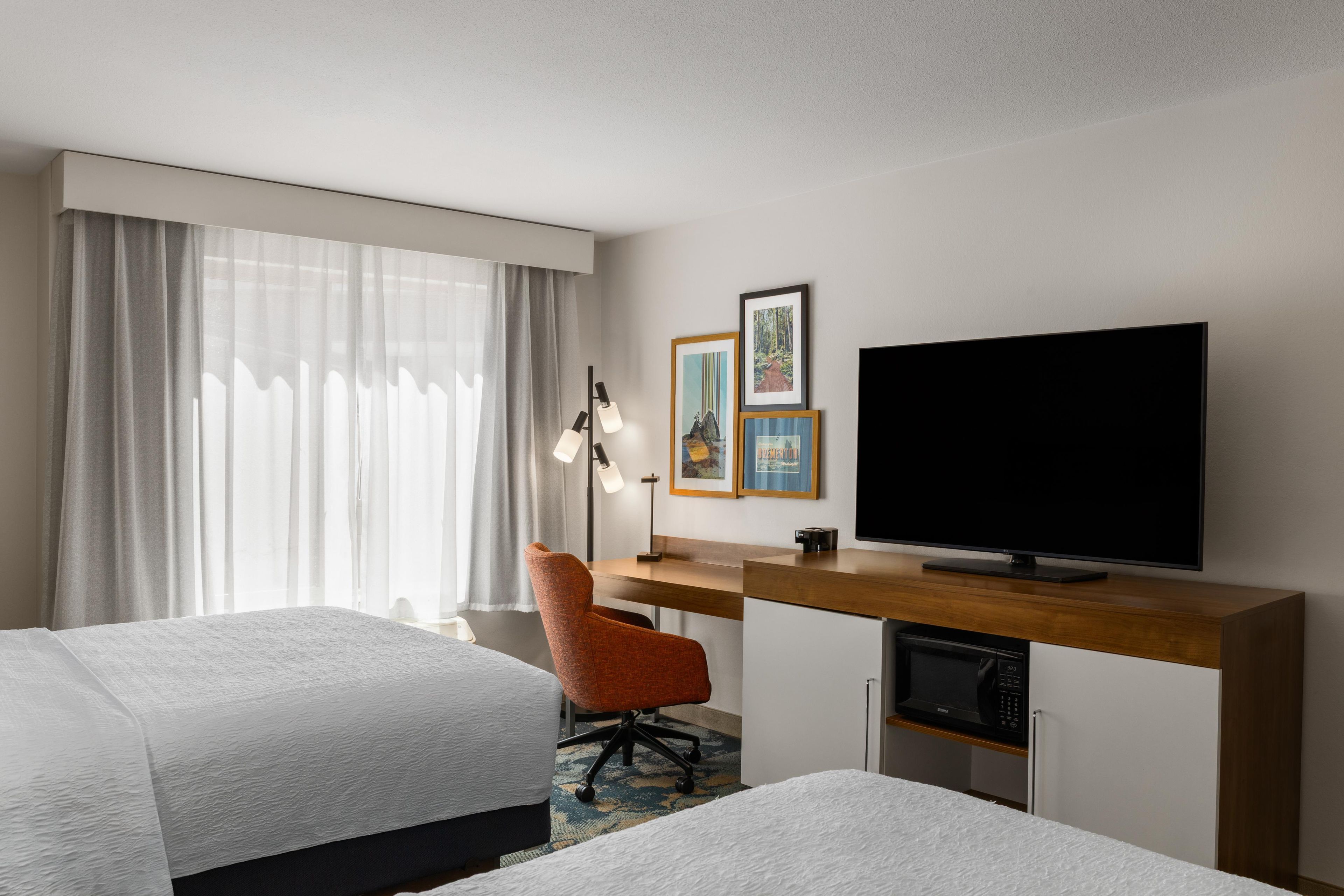 A hotel room with two beds, a desk with a flat-screen TV, a chair, and artwork on the wall.