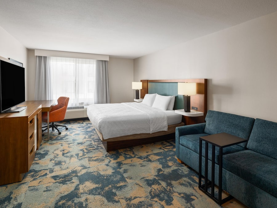 Quarto de hotel com cama, escrivaninha com televisão, sofá e janela com cortinas.