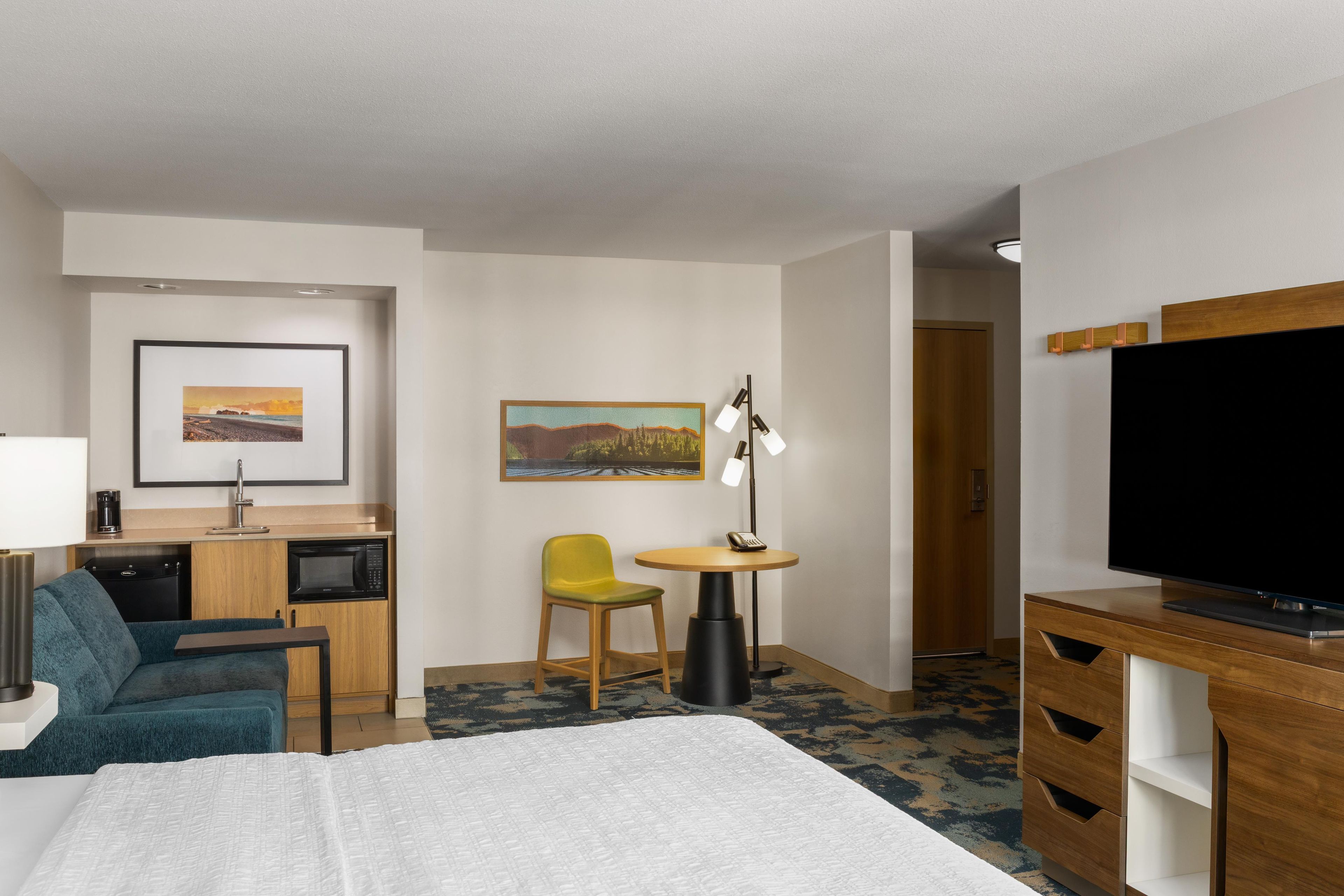 A hotel room with a bed, a desk with a microwave, a chair, a small table with a lamp, a television, and a painting on the wall.