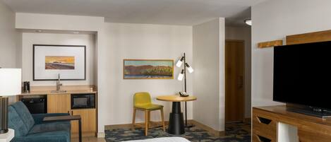 A hotel room with a bed, a desk with a microwave, a chair, a small table with a lamp, a television, and a painting on the wall.