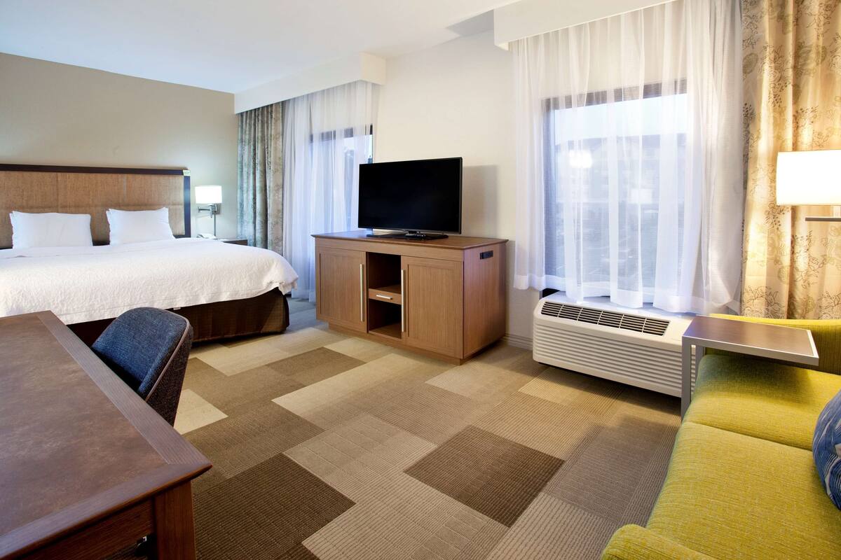 Une chambre d’hôtel avec un lit, un téléviseur posé sur un meuble en bois, un bureau, une chaise, un canapé et une fenêtre avec des rideaux.