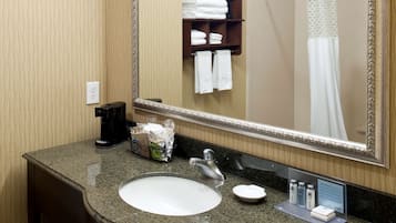 Un baño de hotel con encimera de granito, un lavamanos blanco, un espejo, un perchero para toallas y una cortina de ducha.