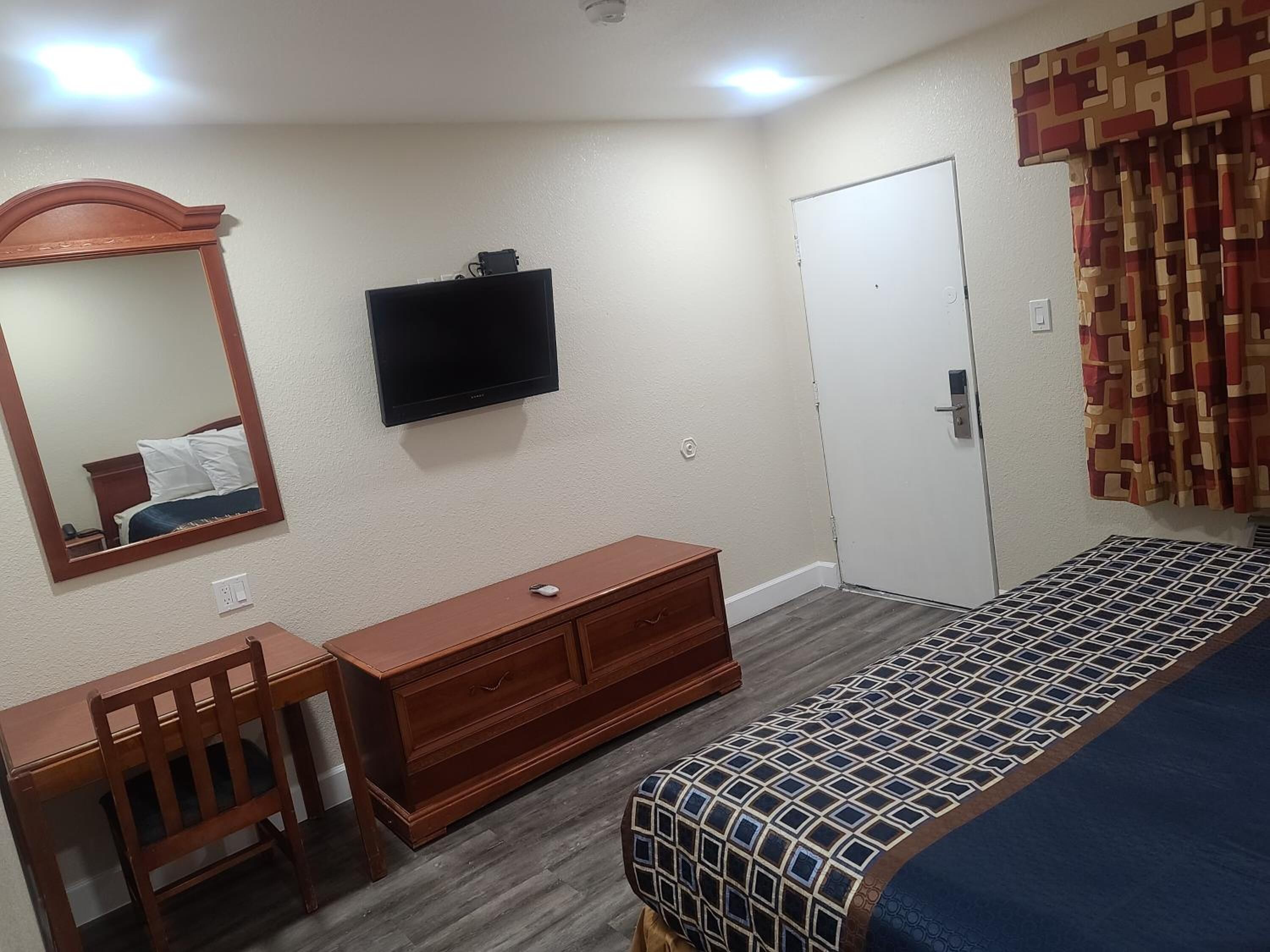 Un dormitorio con cama, cómoda, escritorio, silla, espejo, televisor y puerta.