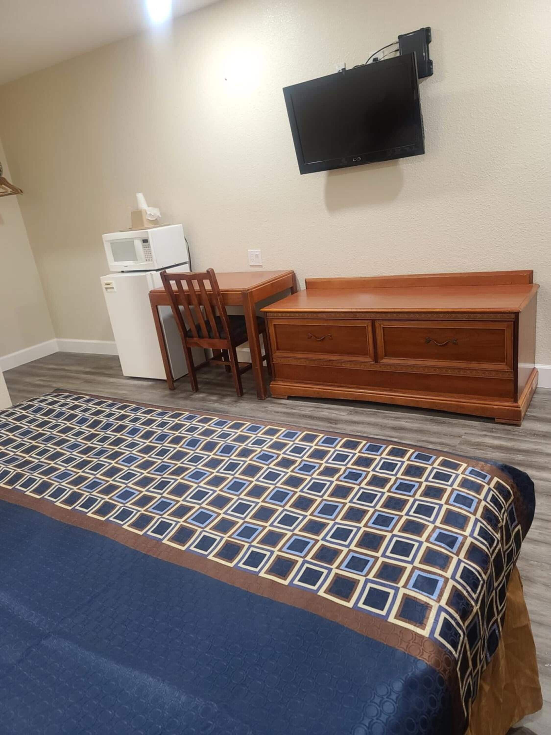 Habitació d'hotel amb llit, una petita còmoda de fusta, escriptori amb cadira, microones i televisió plana penjada a la paret.
