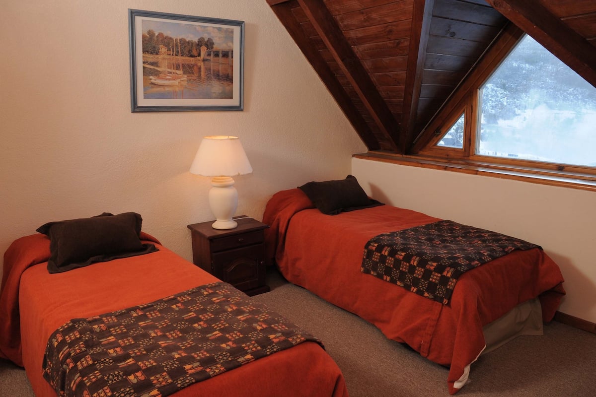 Un dormitorio con dos camas, una mesita de noche con lámpara, un cuadro en la pared y una ventana con vistas a árboles.