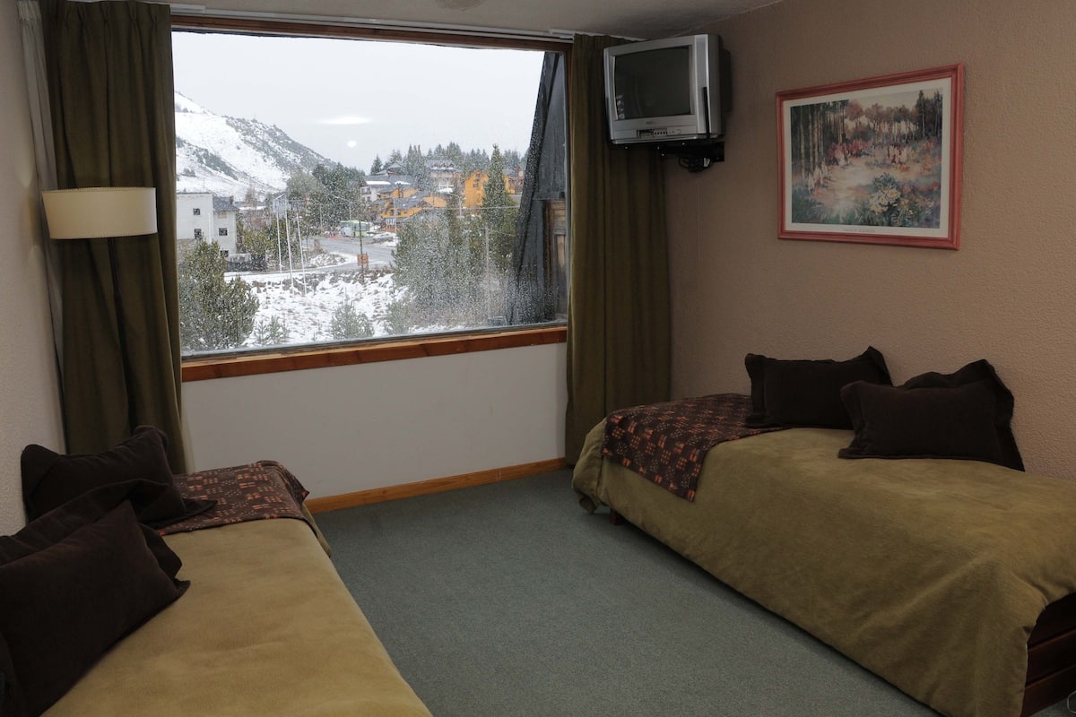 Habitación de hotel con dos camas, una ventana que ofrece vistas a montañas nevadas, un cuadro en la pared y un televisor sobre la ventana.
