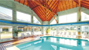 Une piscine intérieure avec des poutres en bois, des chaises longues blanches et de grandes fenêtres.