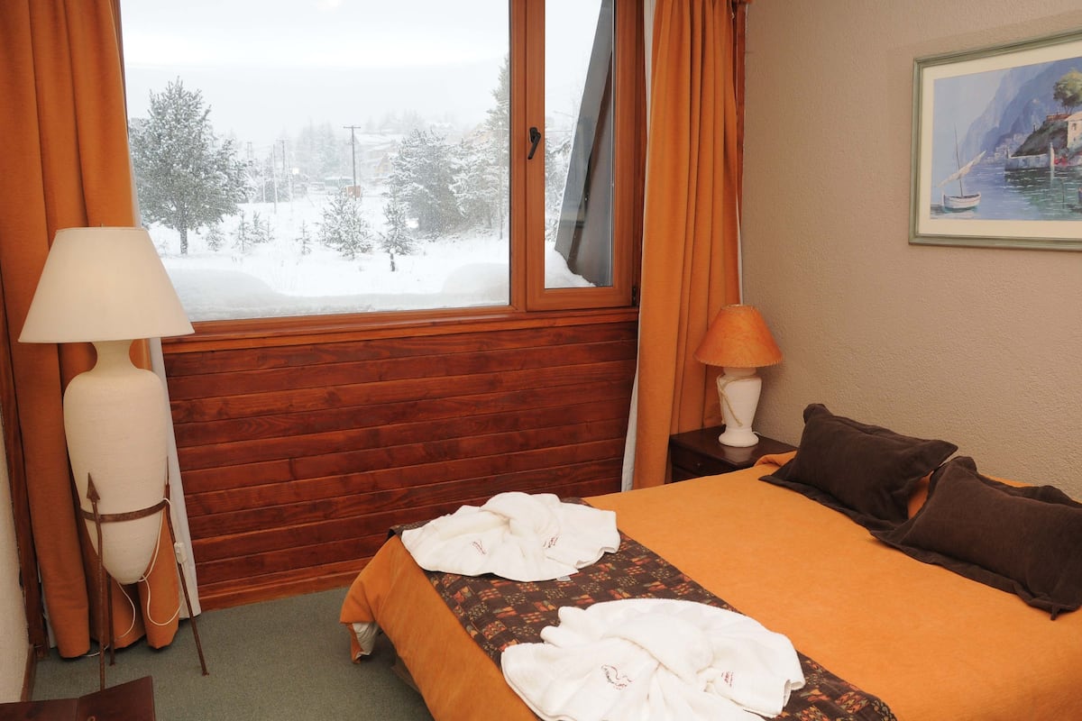 Habitación de hotel con pared de madera, cama con colcha naranja, dos lámparas y un cuadro en la pared.