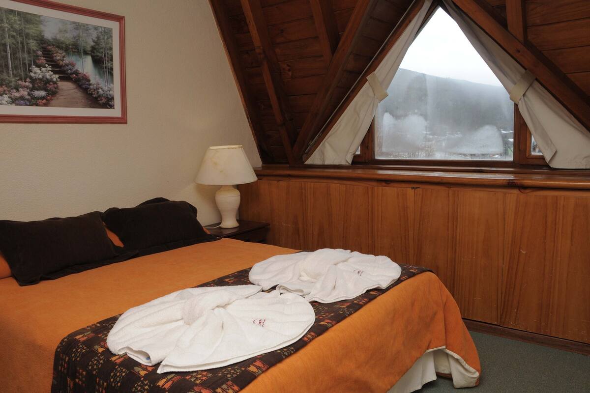 Habitación de hotel con cama, mesita de noche con lámpara, ventana con cortinas e imagen enmarcada en la pared.
