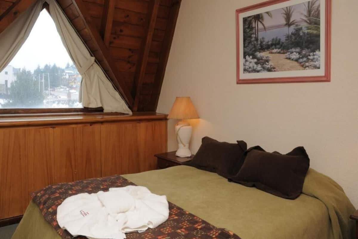 Una habitación con cama, mesita de noche con lámpara, ventana con cortinas e imagen enmarcada en la pared.