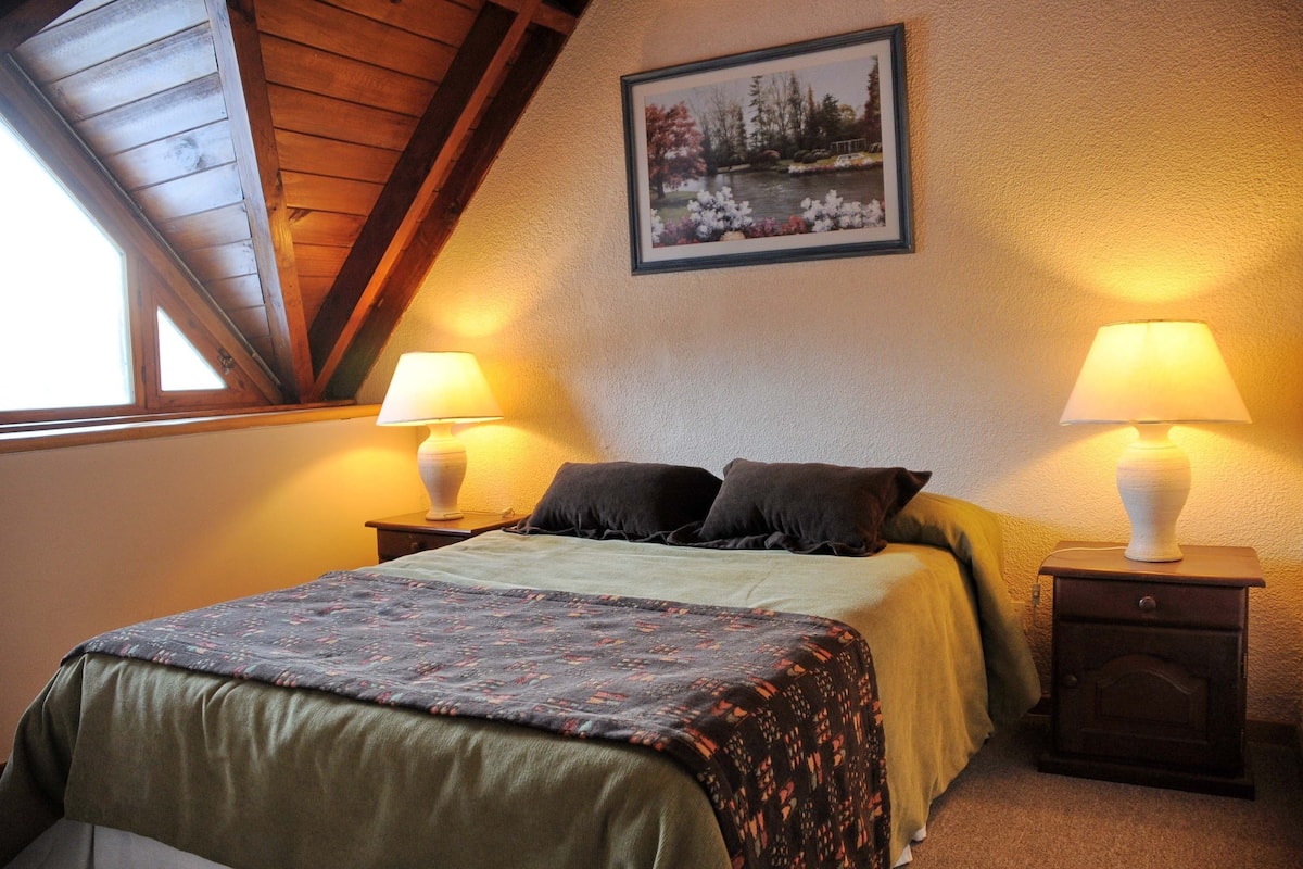 Un dormitorio con cama, dos mesitas de noche con lámparas, un cuadro enmarcado en la pared y un techo inclinado.