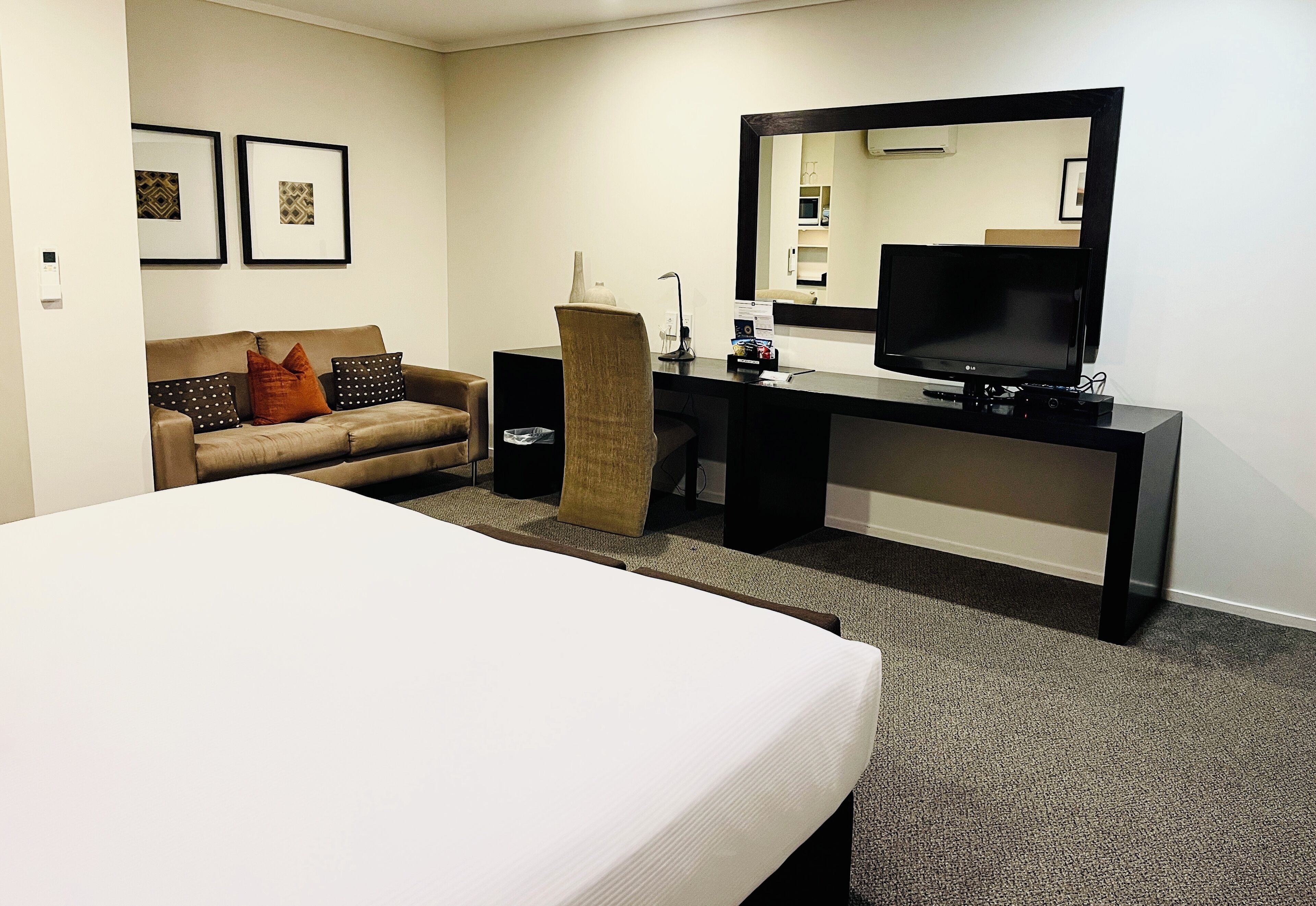 A hotel room with a bed, a desk with a flat-screen TV, a chair, a sofa, and two framed pictures on the wall.