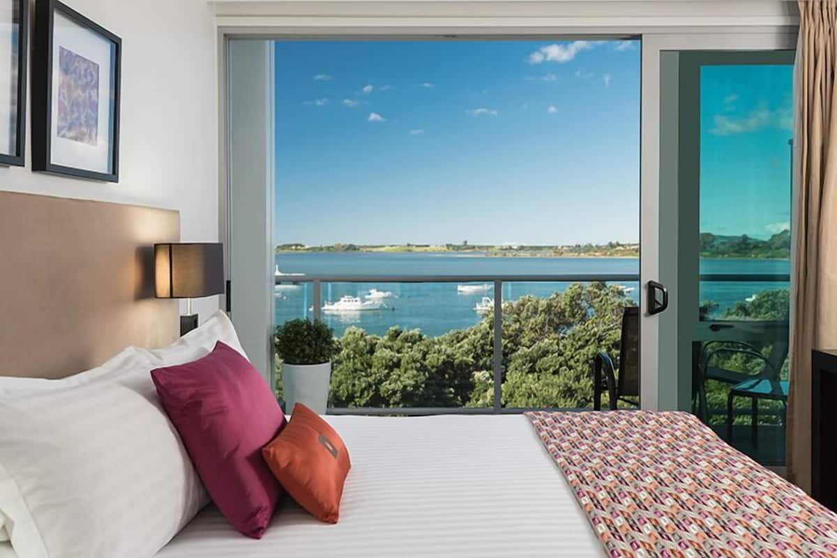 A hotel room with a large window offering a view of a body of water and boats, a bed with pillows, a small potted plant, and a chair by the window.