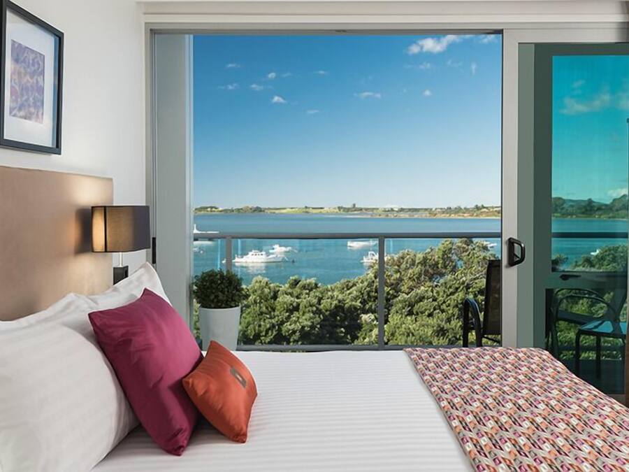 Habitación de hotel con un ventanal que ofrece vistas a un cuerpo de agua y barcos, una cama con almohadas, una pequeña planta en maceta y una silla junto a la ventana.