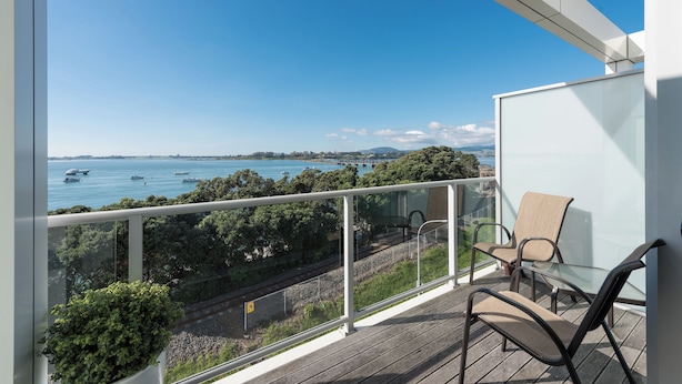 A balcony with a glass railing overlooking a body of water with boats, greenery, and a distant shoreline. - Hotel on Devonport (Tauranga)
