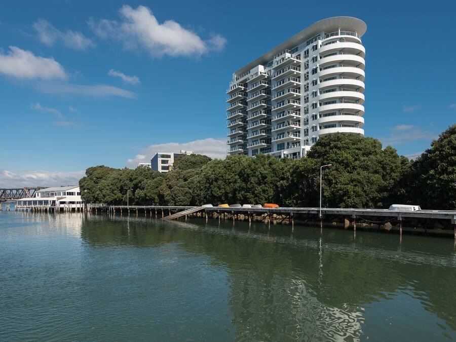 A modern high-rise hotel with multiple balconies, situated near a body of water with a bridge in the background.