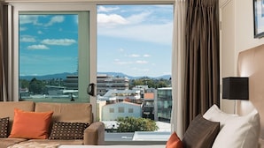 A hotel room with a large sliding glass door leading to a balcony, a sofa with cushions, a bed, and a nightstand with a lamp. - Hotel on Devonport (Tauranga)