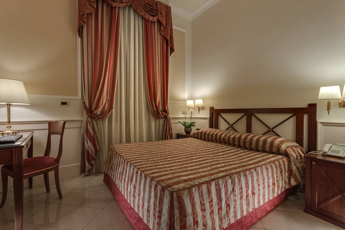 A hotel room with a large bed, a desk with a chair, a telephone, and a nightstand with a lamp.