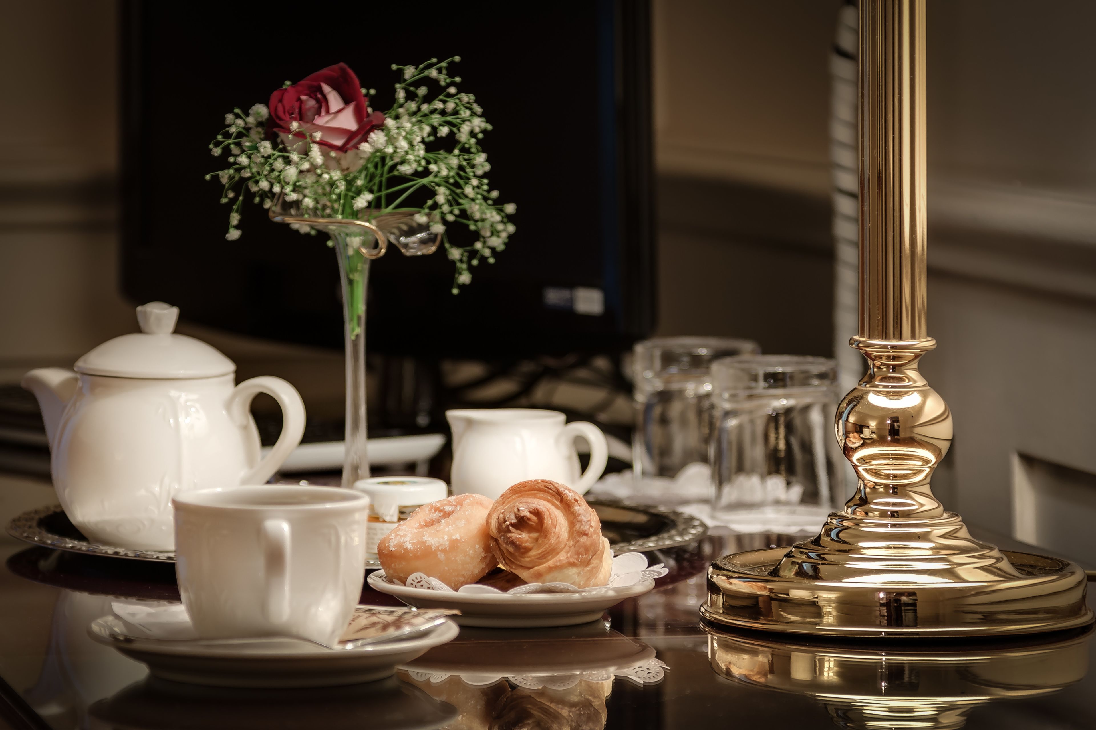 A table with a teapot, cups, pastries, a vase with flowers, and a lamp.