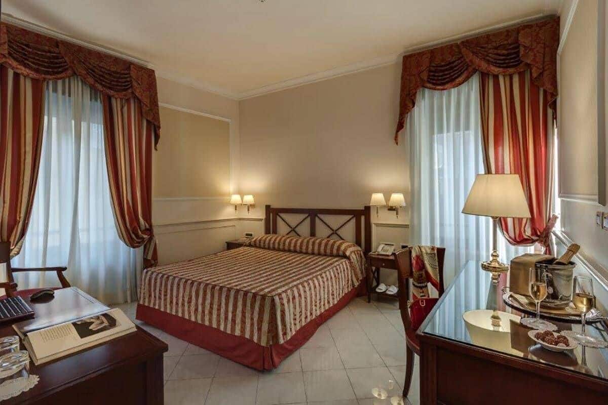 A hotel room with a large bed, two bedside tables with lamps, a desk with a chair, and a small table with a lamp and a bottle.