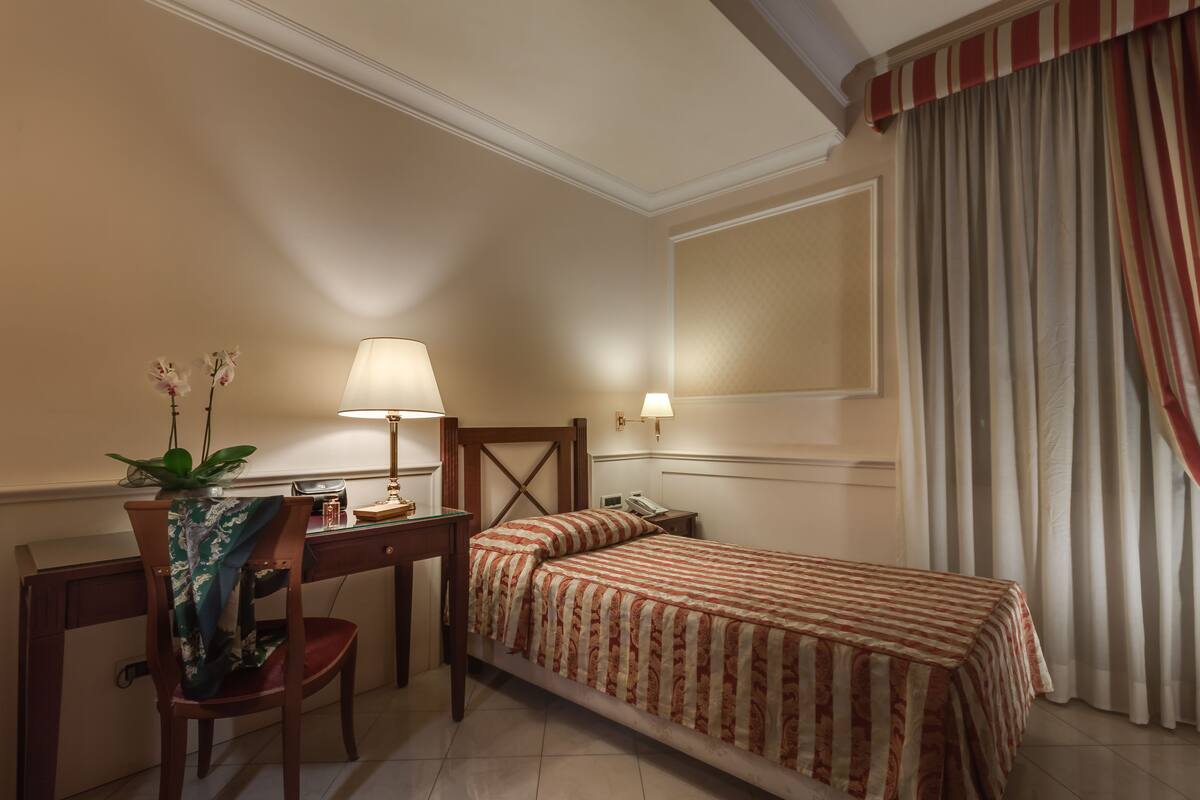 A hotel room with a bed, desk, chair, lamp, and a potted plant.