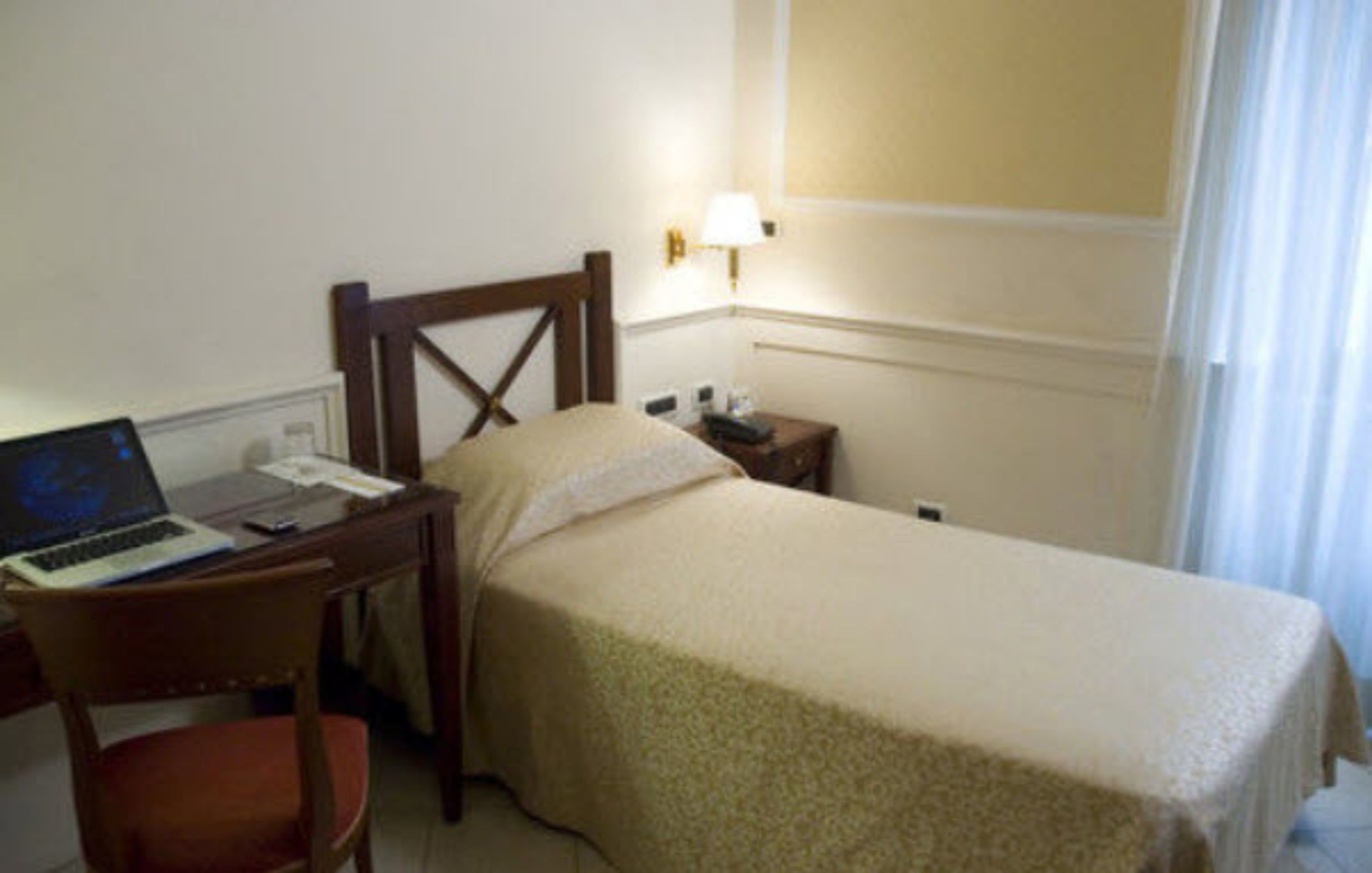 Habitación de hotel con cama, escritorio con una computadora portátil, silla, mesita de noche y lámpara de pared.