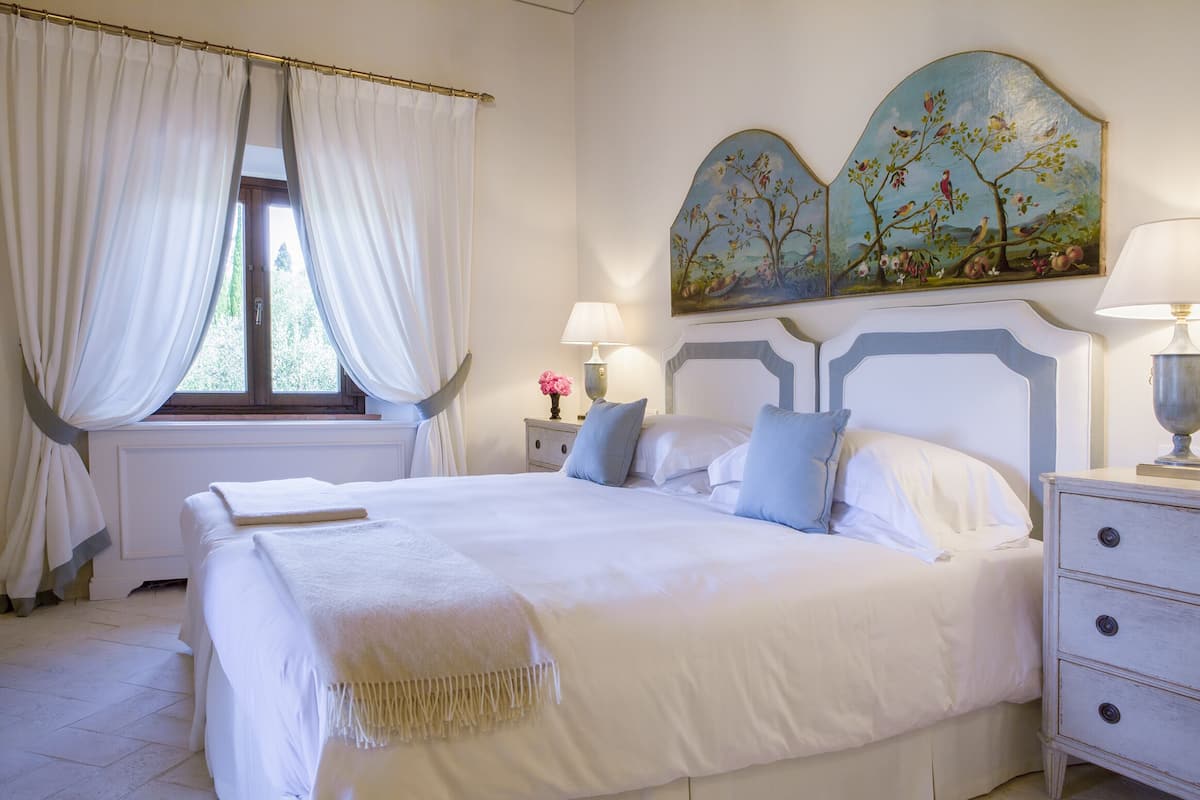 Une chambre à coucher avec un grand lit, du linge de lit blanc, une table de chevet avec une lampe, un tableau au-dessus du lit et une fenêtre avec des rideaux.
