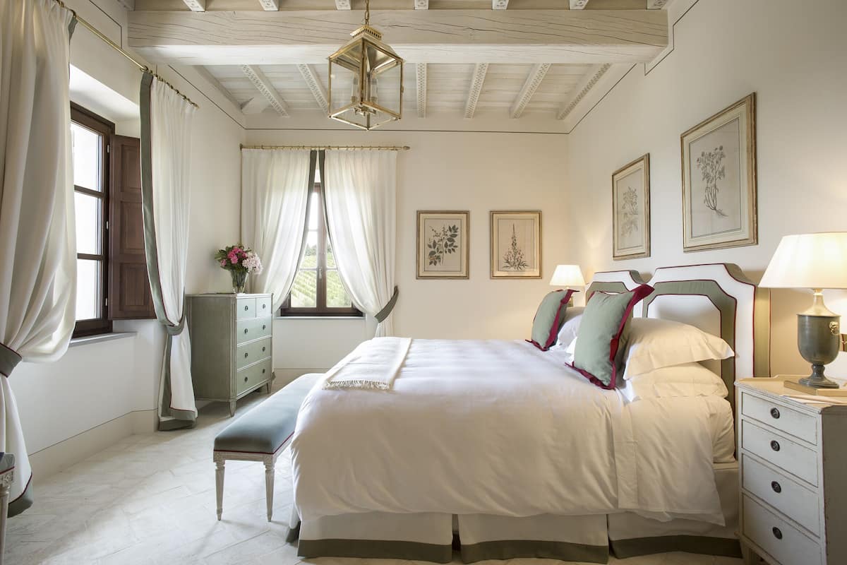 Une chambre à coucher avec un grand lit, des tables de chevet, un banc, une commode et des œuvres d’art encadrées accrochées aux murs.