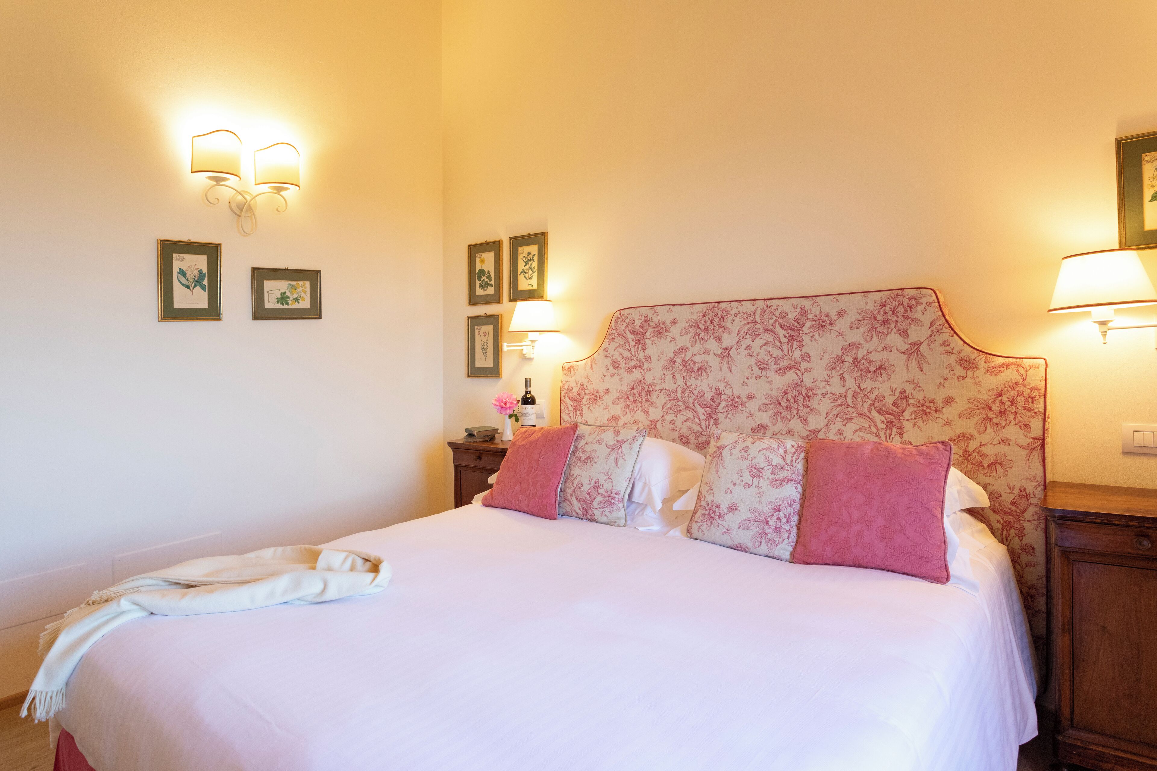 A neatly made bed with floral headboard, bedside lamps, and framed pictures on the wall.
