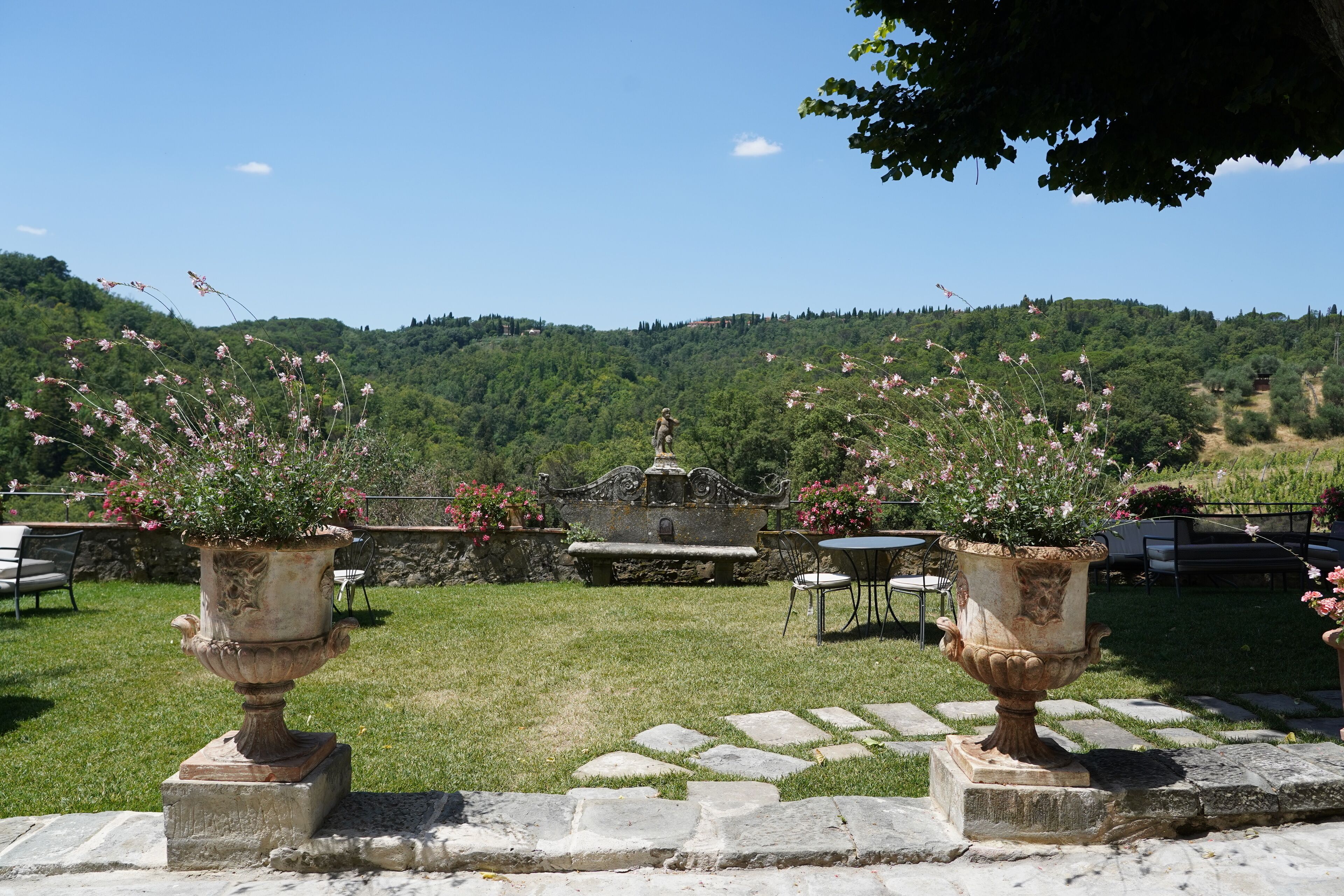 Un jardin agrémenté de deux grands bacs en pierre ornés de fleurs roses, d’un banc en pierre et d’une table avec des chaises.