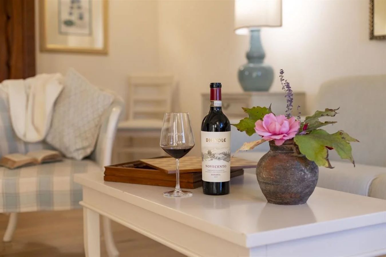 A bottle of Novecento wine, a glass of wine, and a vase with flowers on a white table in a living room.