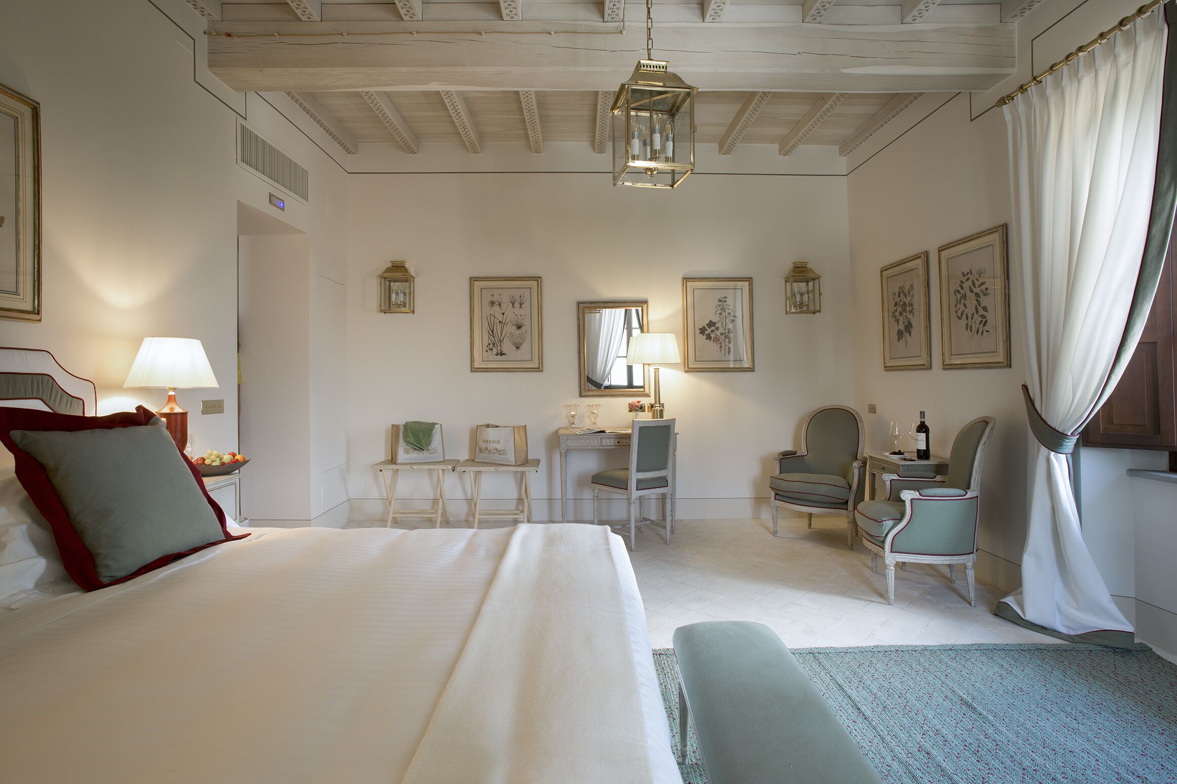Habitación de hotel con una cama grande, un escritorio, dos sillas, una mesita con una botella y cuadros en las paredes.