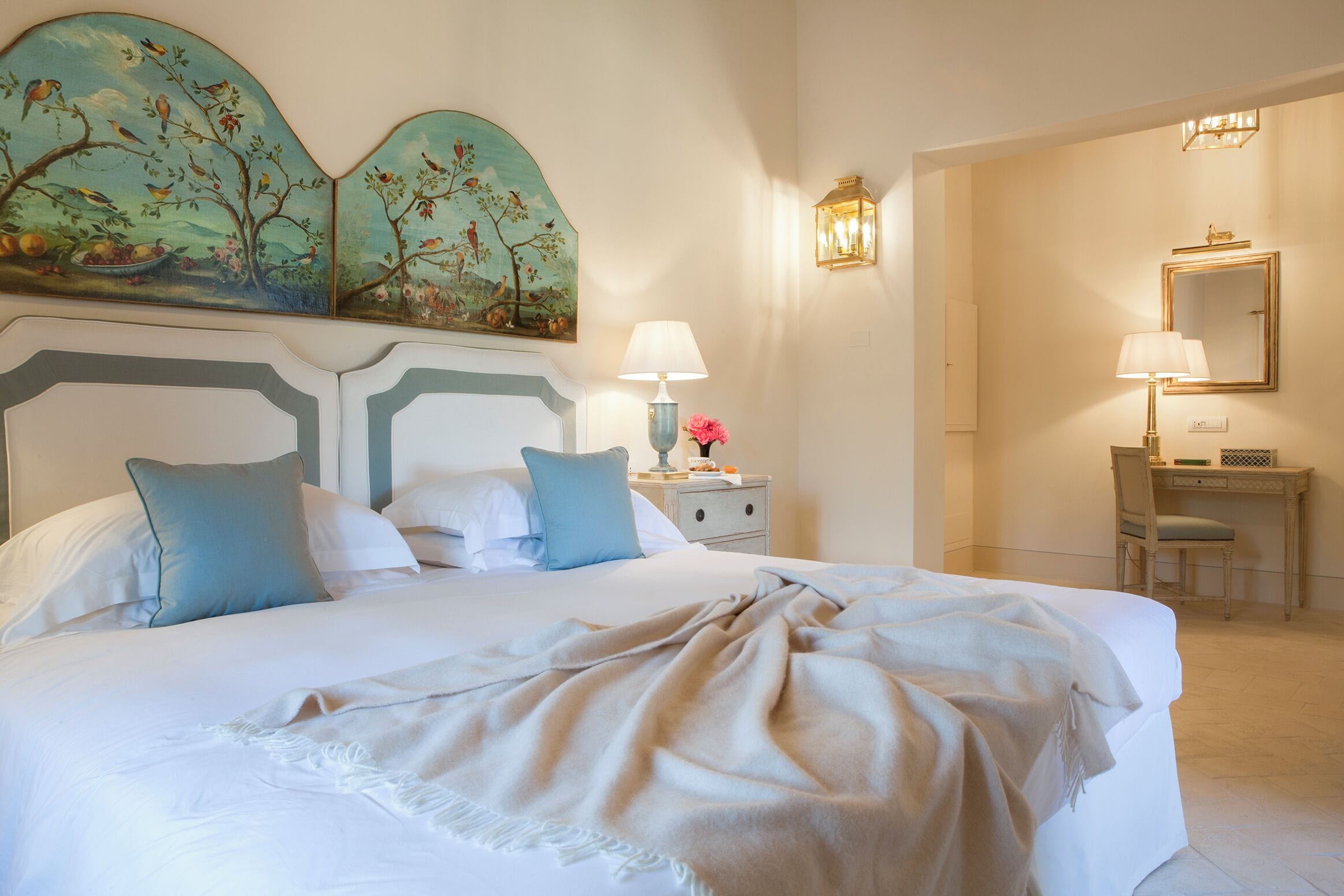 A neatly made bed with a white and beige duvet, a nightstand with a lamp, and a desk with a chair.