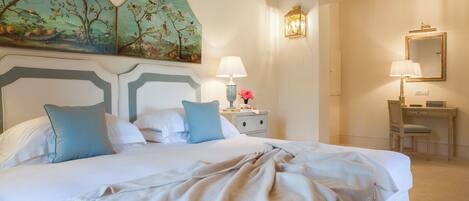 A neatly made bed with a white and beige duvet, a nightstand with a lamp, and a desk with a chair.