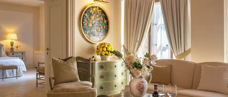 A well-lit living room with a sofa, a coffee table with a vase and wine glasses, a painting on the wall, and a bed in the background.