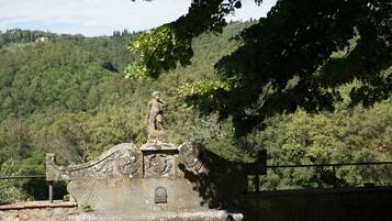 Un banco de piedra con una estatua encima, rodeado de follaje y colinas al fondo.
