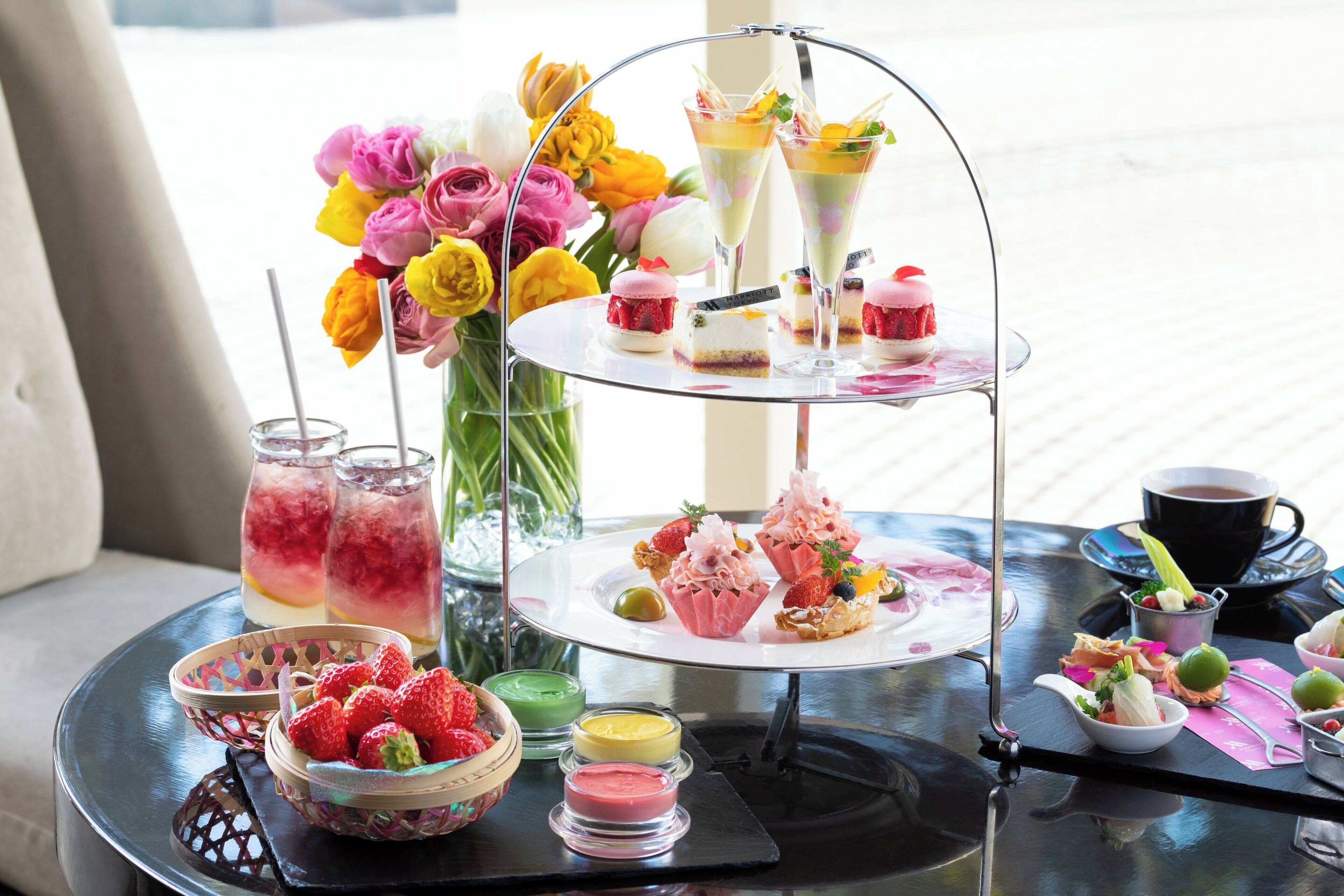 Een drijvend taartplateau met verschillende gebakjes en drankjes, een schaal aardbeien en een vaas bloemen op een tafel.
