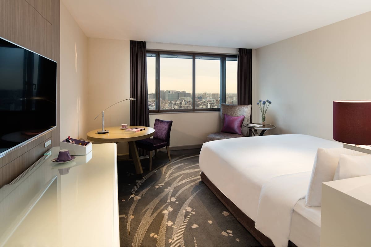A hotel room with a bed, a desk, a TV, a window with a city view, a small table, and a chair.
