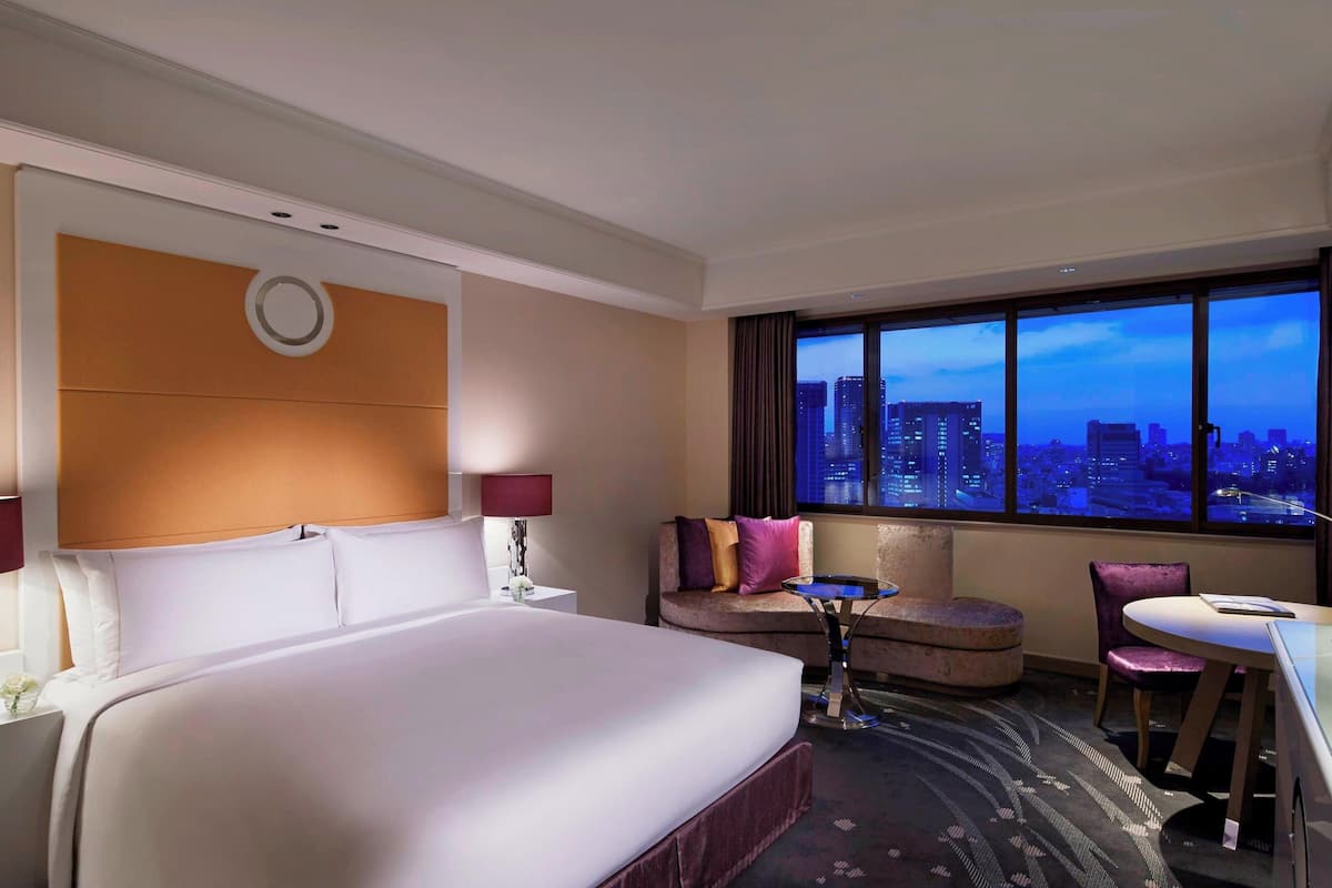 A hotel room with a large bed, a seating area with a table and chairs, and a view of a cityscape through large windows.