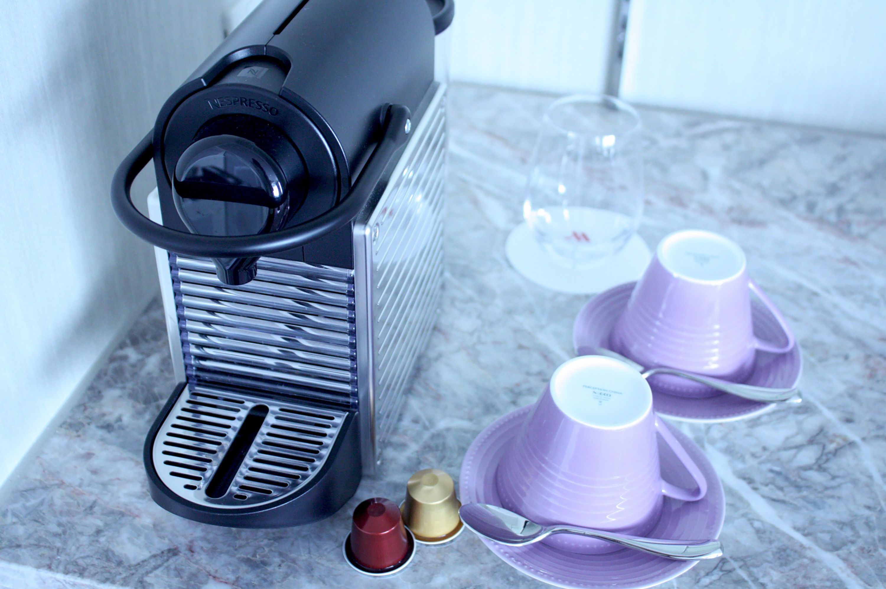 Una cafetera con depósito de agua, dos tazas moradas con sus platillos y un vaso de agua sobre una encimera de mármol.