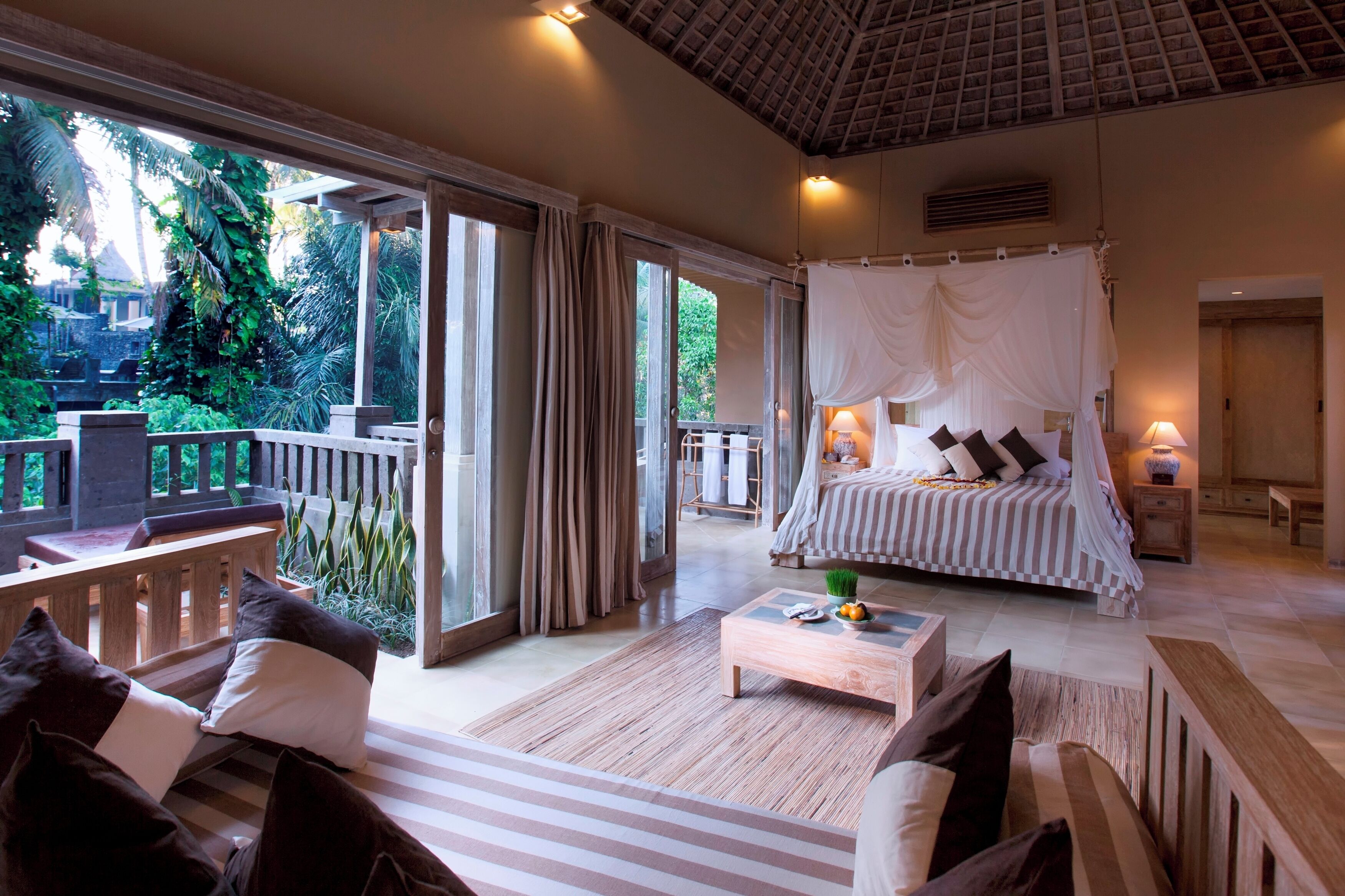 A bedroom with a canopy bed, a wooden bench, a coffee table, and a view of the garden.