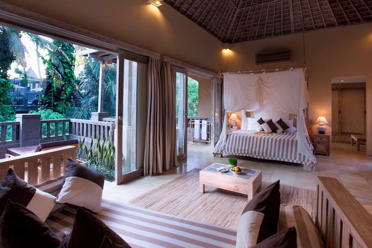 A bedroom with a canopy bed, a wooden bench, a coffee table, and a view of the garden.