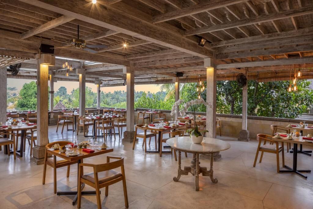 An open-air restaurant with wooden tables and chairs, large windows, and a view of greenery.