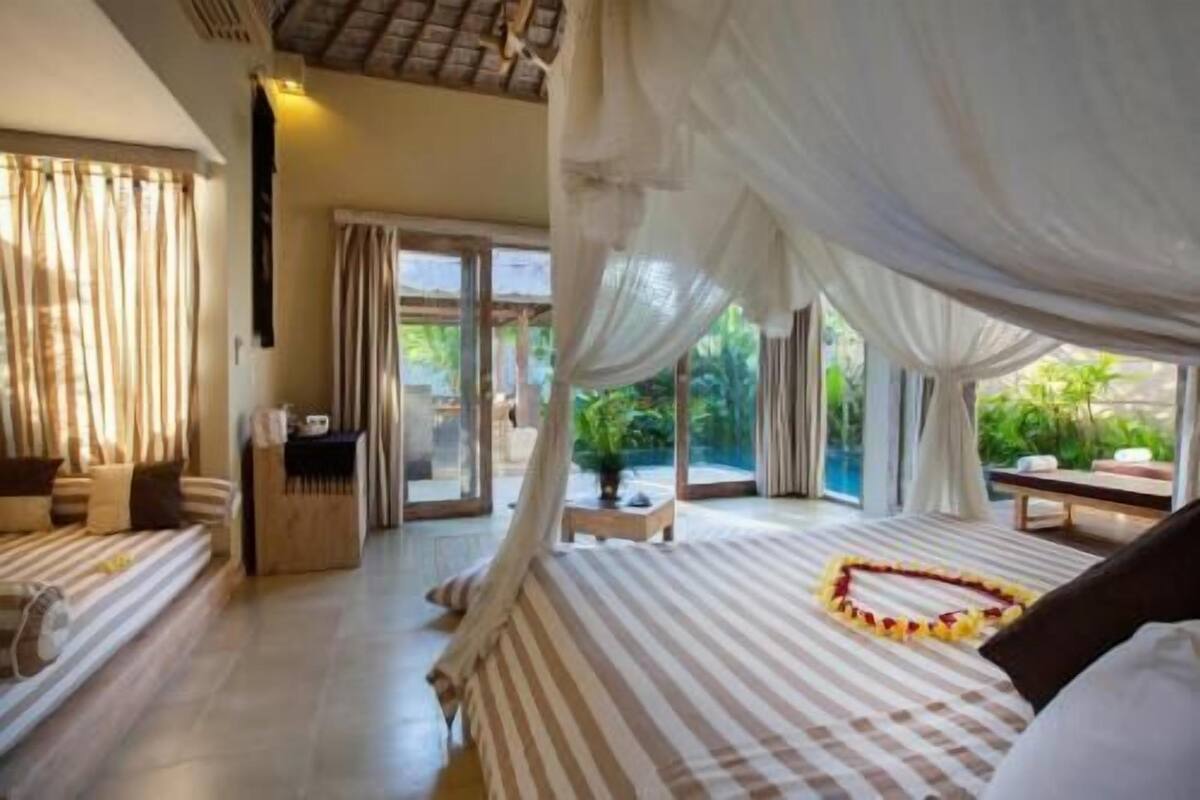 A bedroom with a canopy bed, a view of greenery outside, and a small table with a plant.