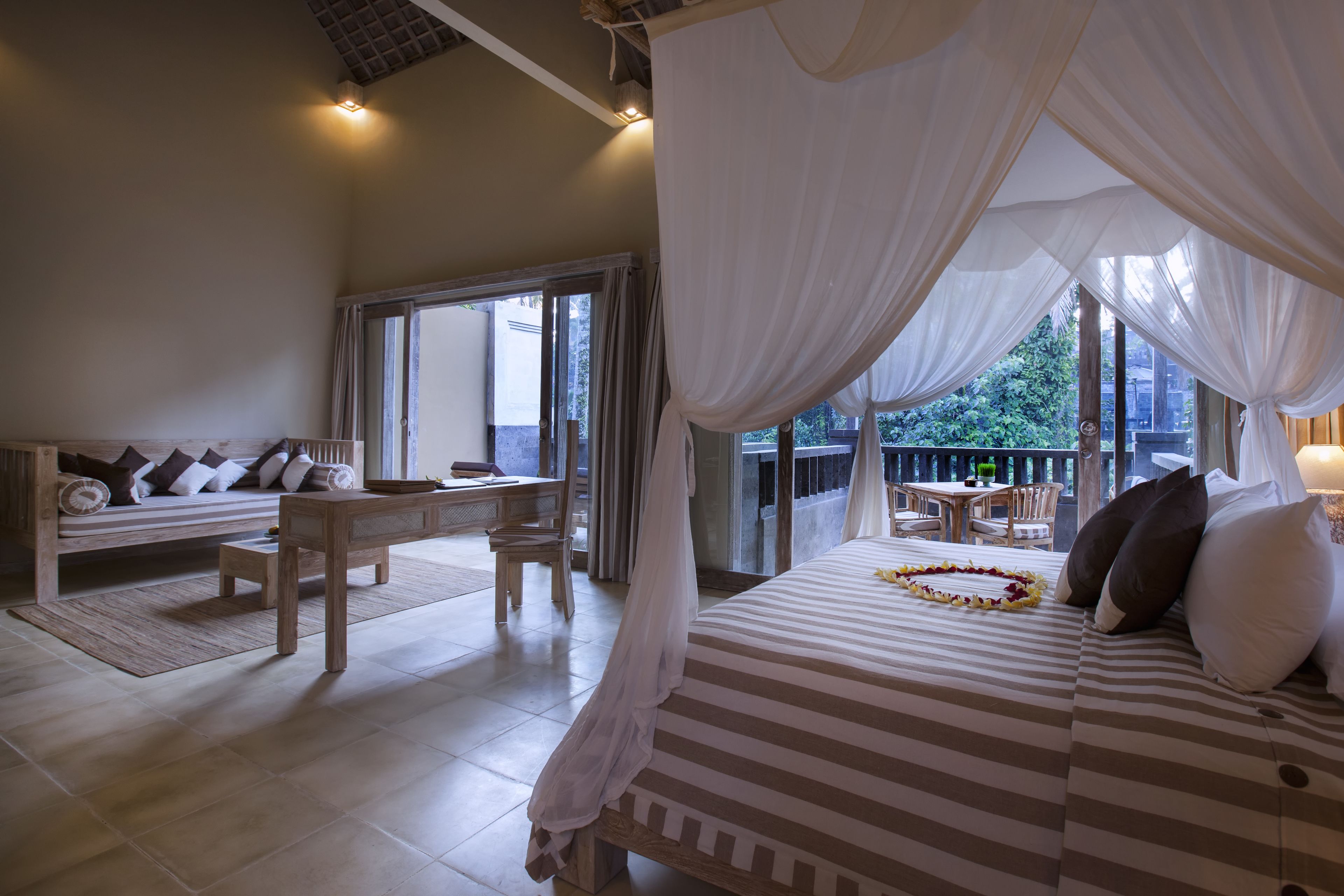 A bedroom with a bed, a sitting area with a table and chairs, and a balcony with a view of greenery.