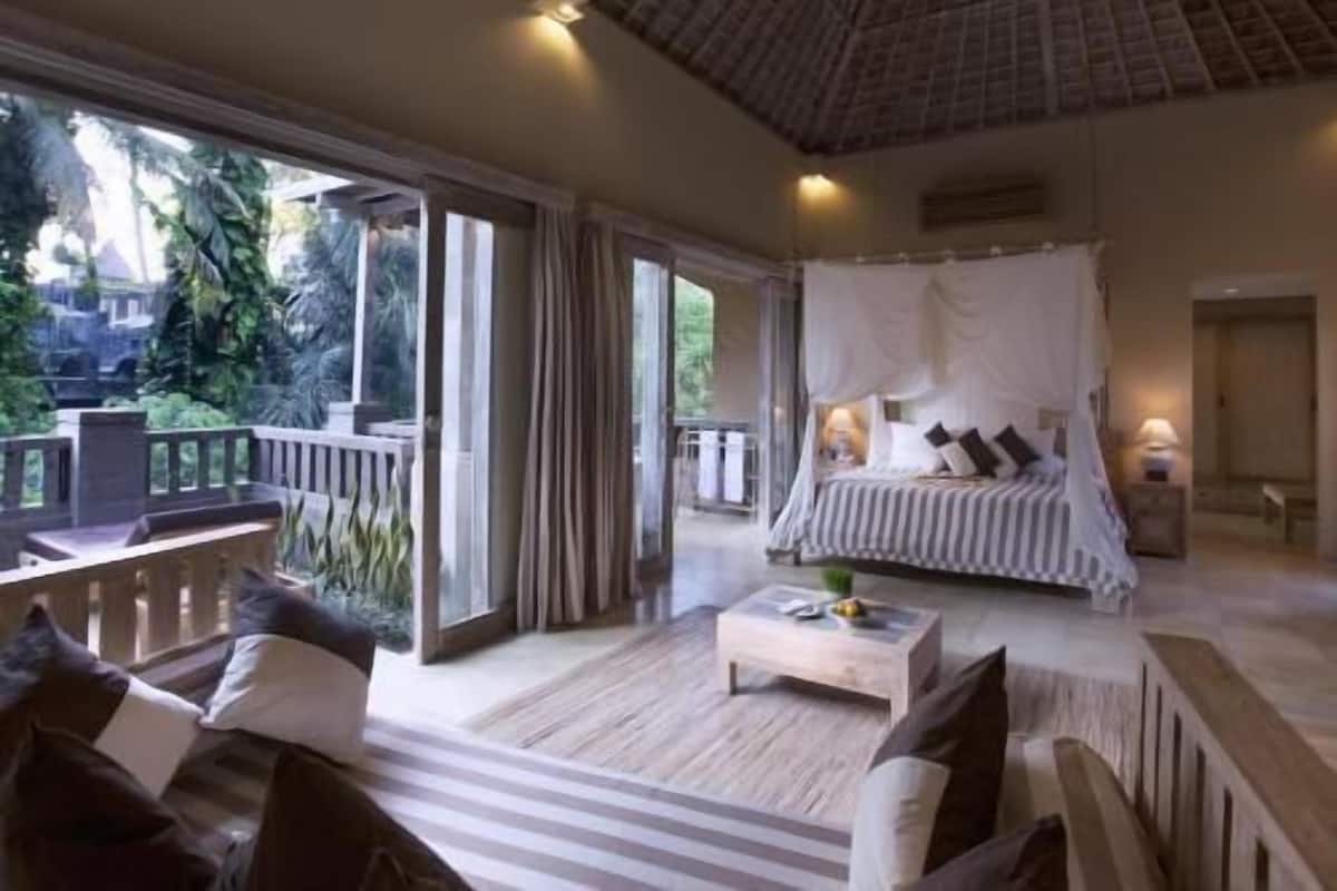 A spacious bedroom with a large bed, a sitting area with a coffee table, and a balcony with a view of the surrounding greenery.