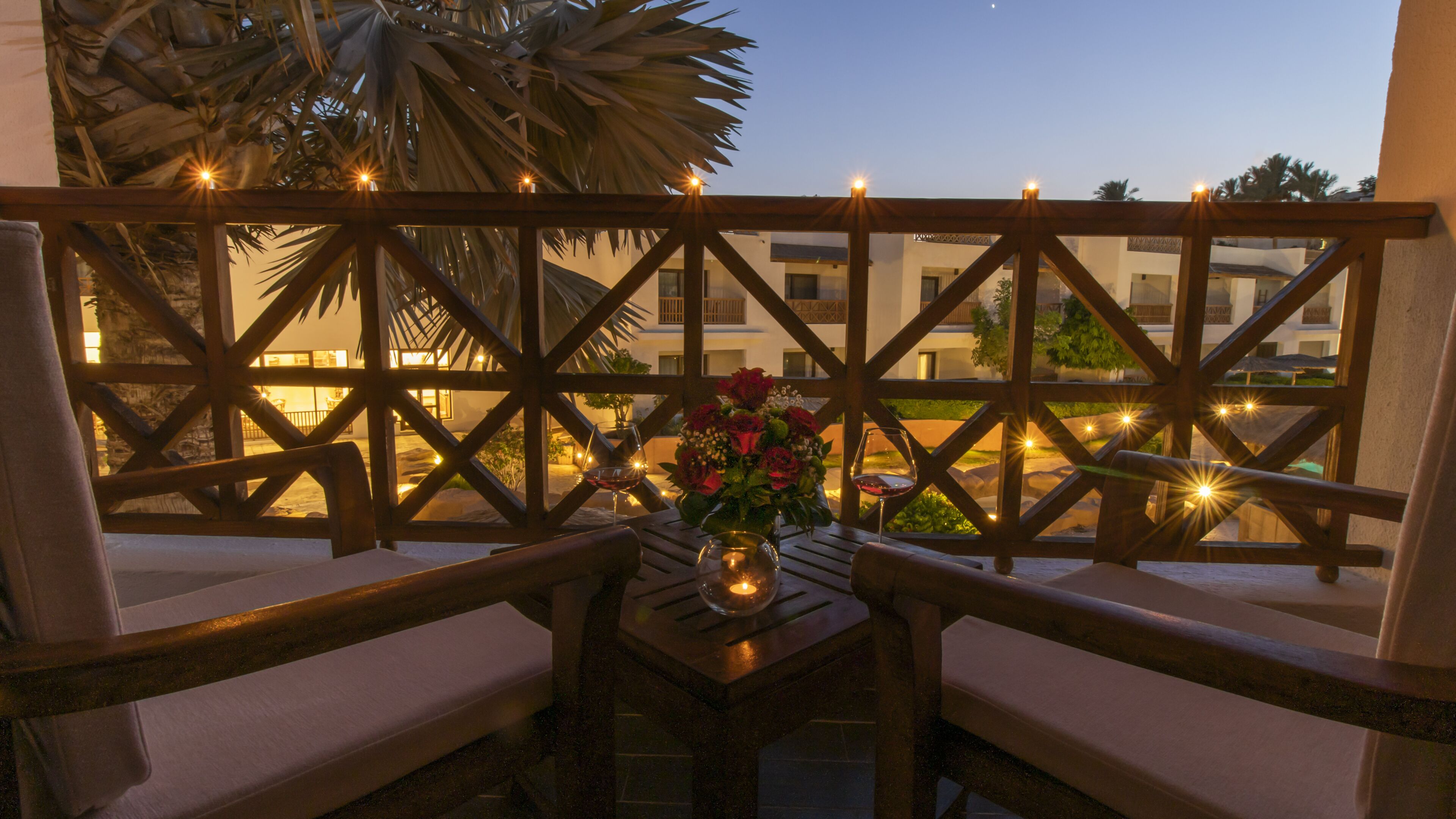 A balcony with wooden lattice, two cushioned chairs, a table with a candle and a vase of flowers, and a view of buildings in the background.