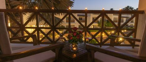 A balcony with wooden lattice, two cushioned chairs, a table with a candle and a vase of flowers, and a view of buildings in the background.